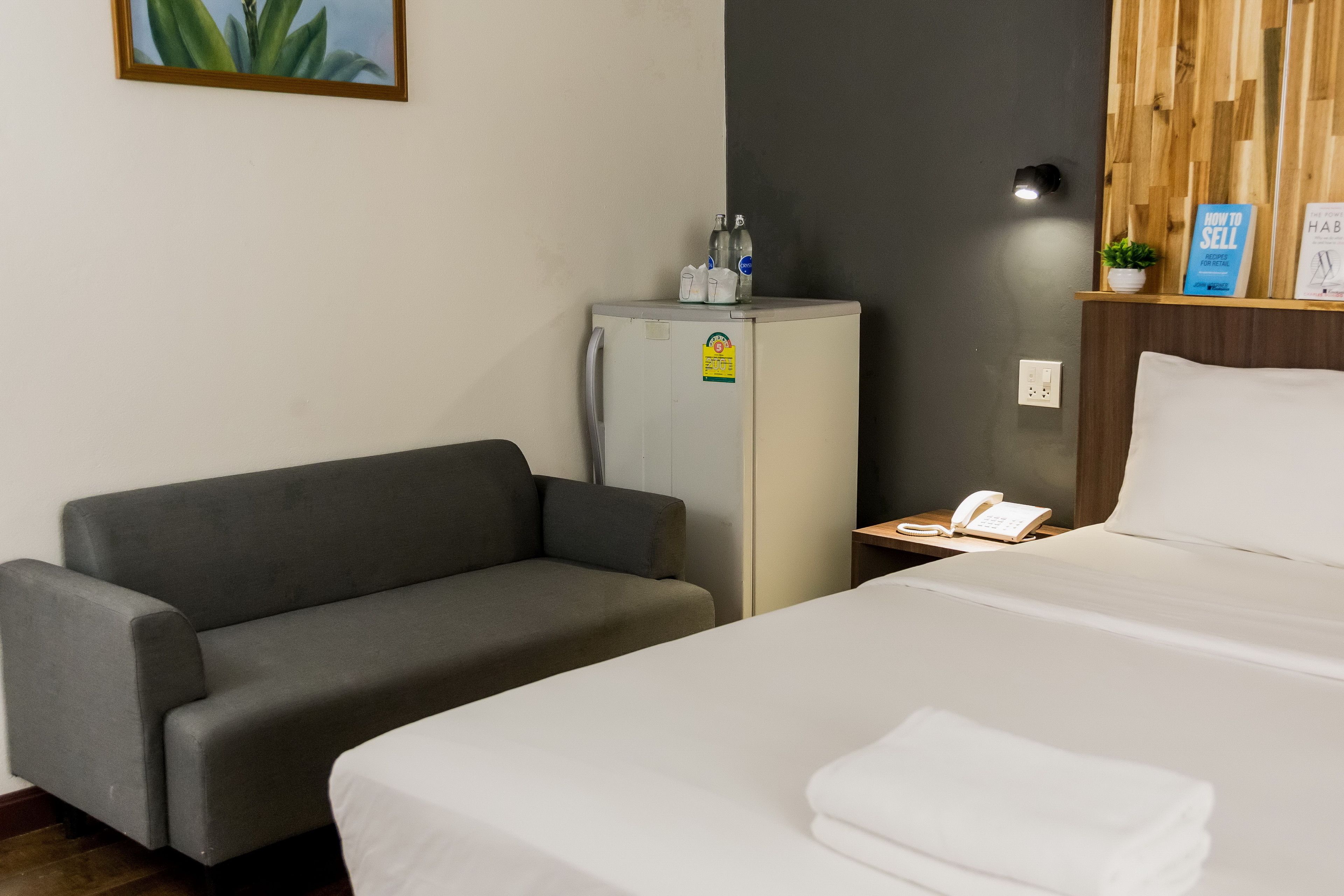 A hotel room with a bed, a grey sofa, a small refrigerator, a bedside table with a phone, and a wall-mounted shelf with books and a plant.