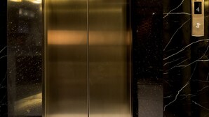 Um elevador com acabamento metálico, em frente a uma parede de mármore preto com veios brancos.