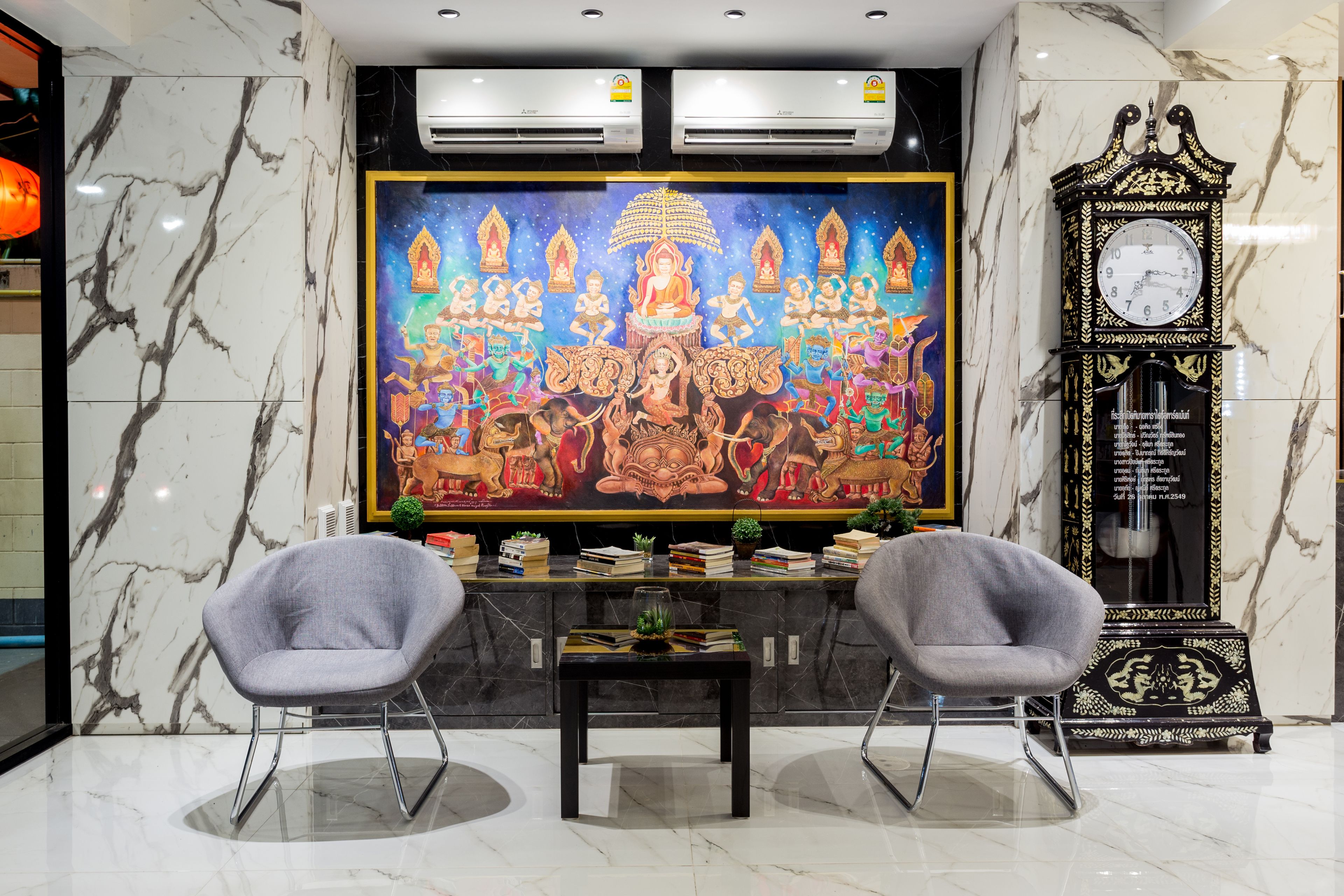 A lobby with marble walls, a large mural, two grey chairs, a small table, and a traditional clock.