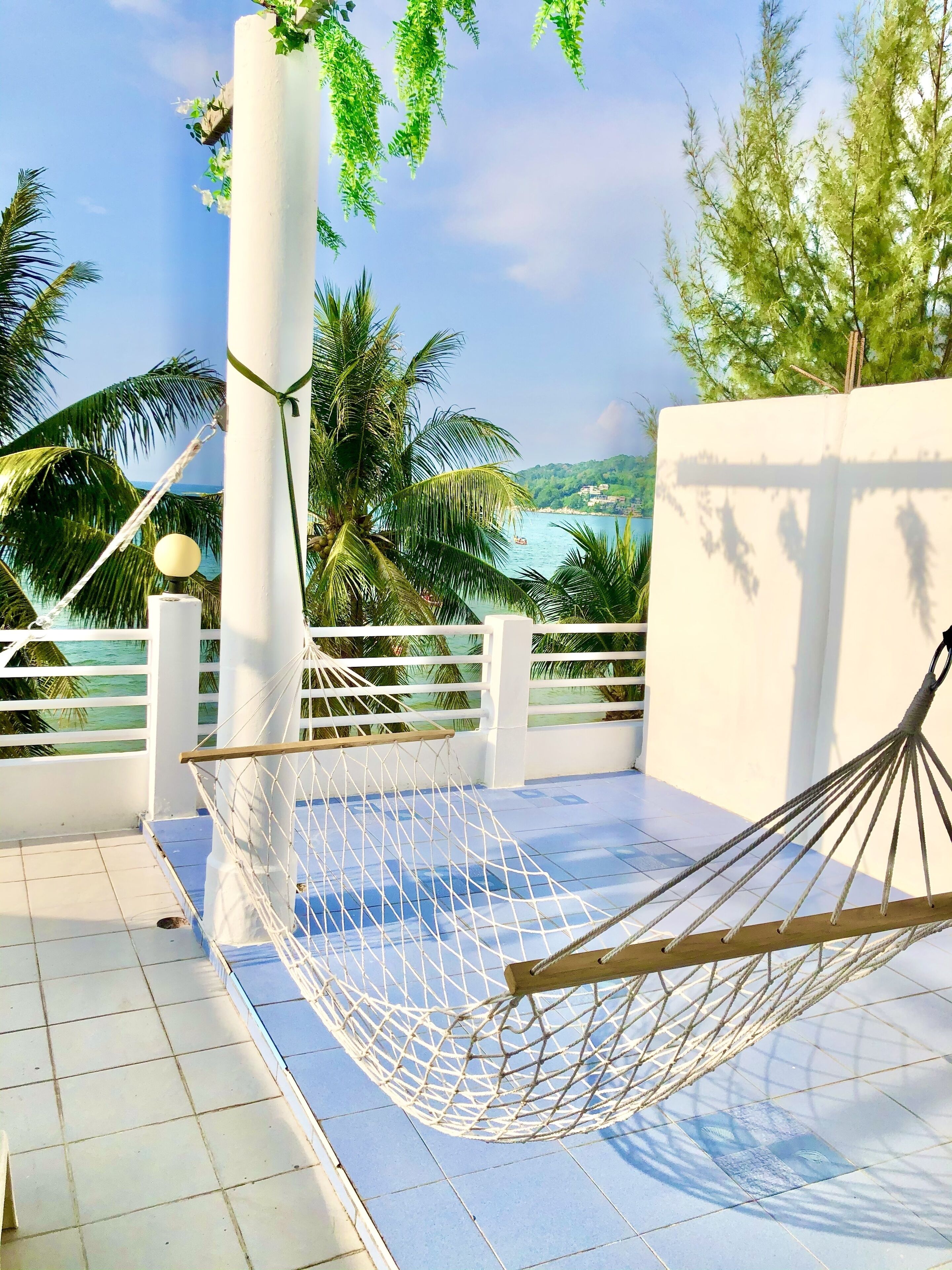 A hammock on a balcony with a view of the sea and palm trees.