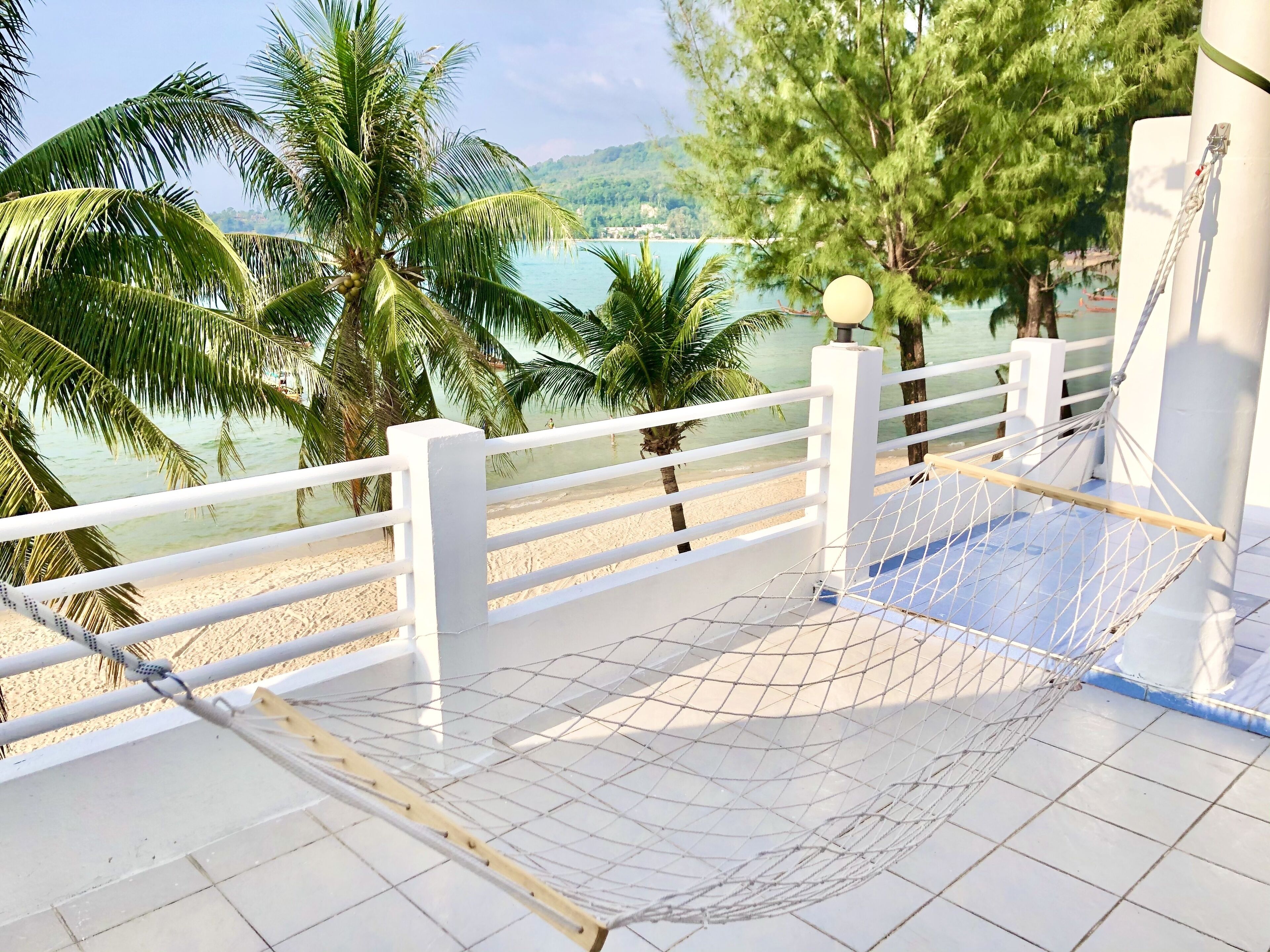 A hammock on a balcony with a view of a beach and palm trees.