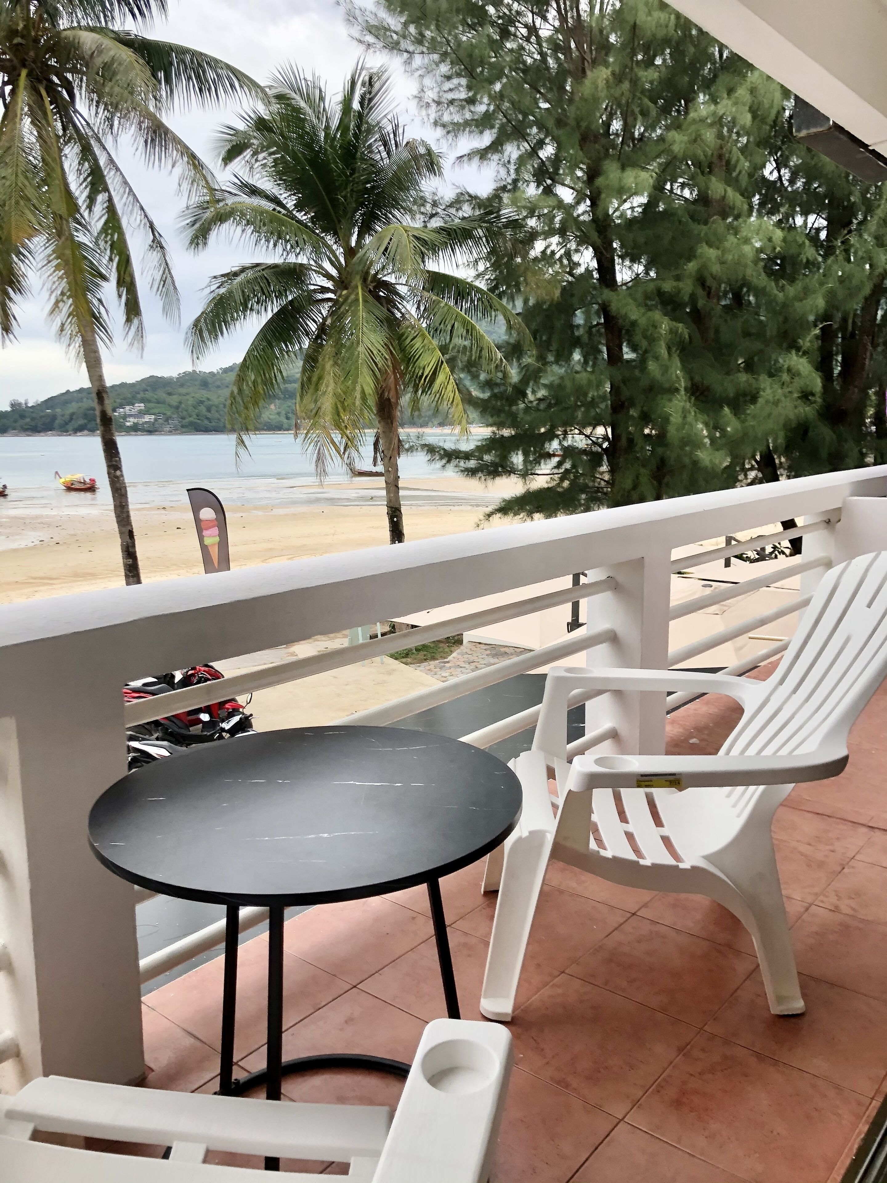 A balcony with a table and chairs, overlooking a beach with palm trees and a view of the sea.
