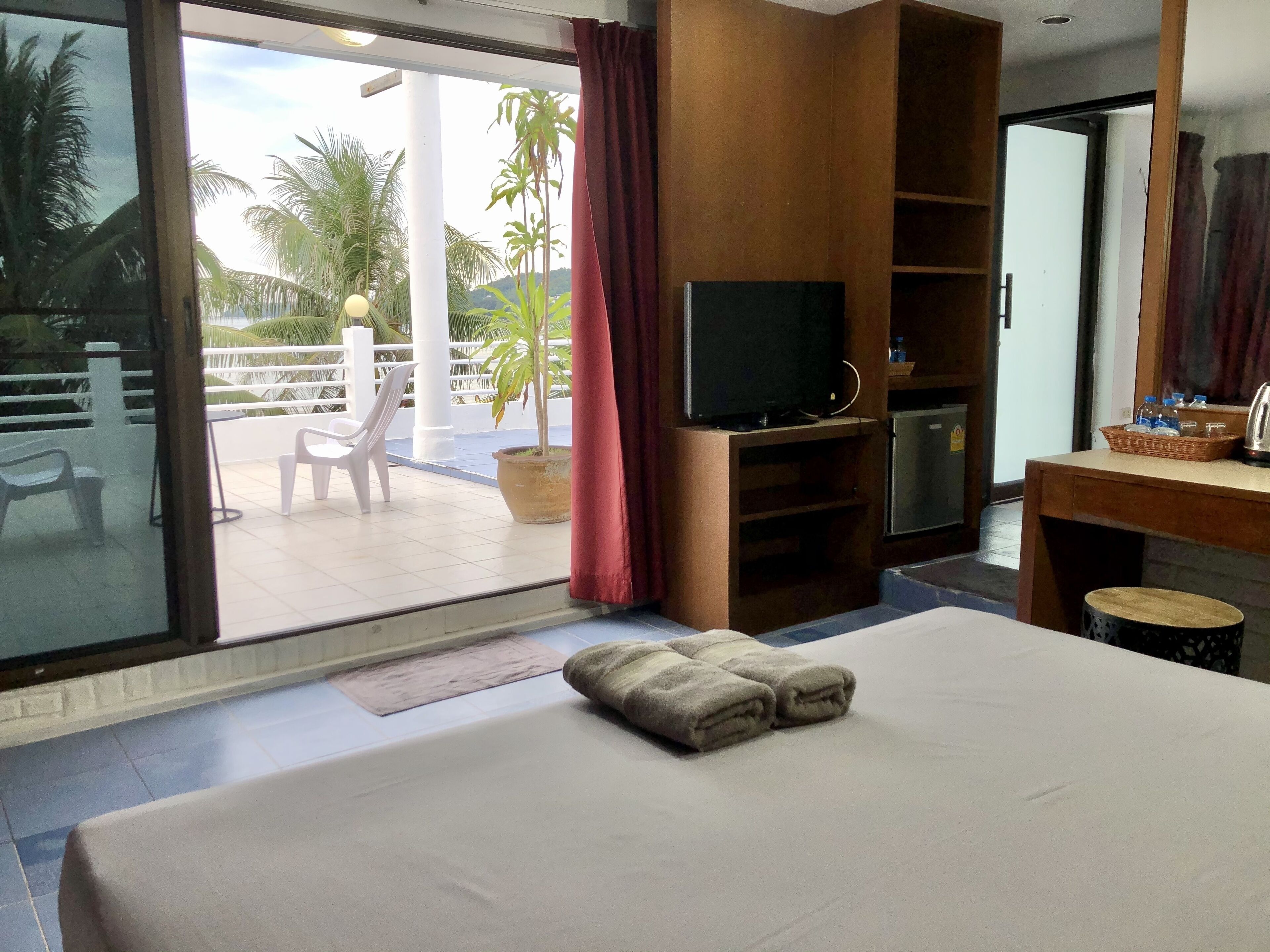 A hotel room with a large bed, a TV, a desk with a basket of bottled water, and a view of palm trees through the sliding glass door.