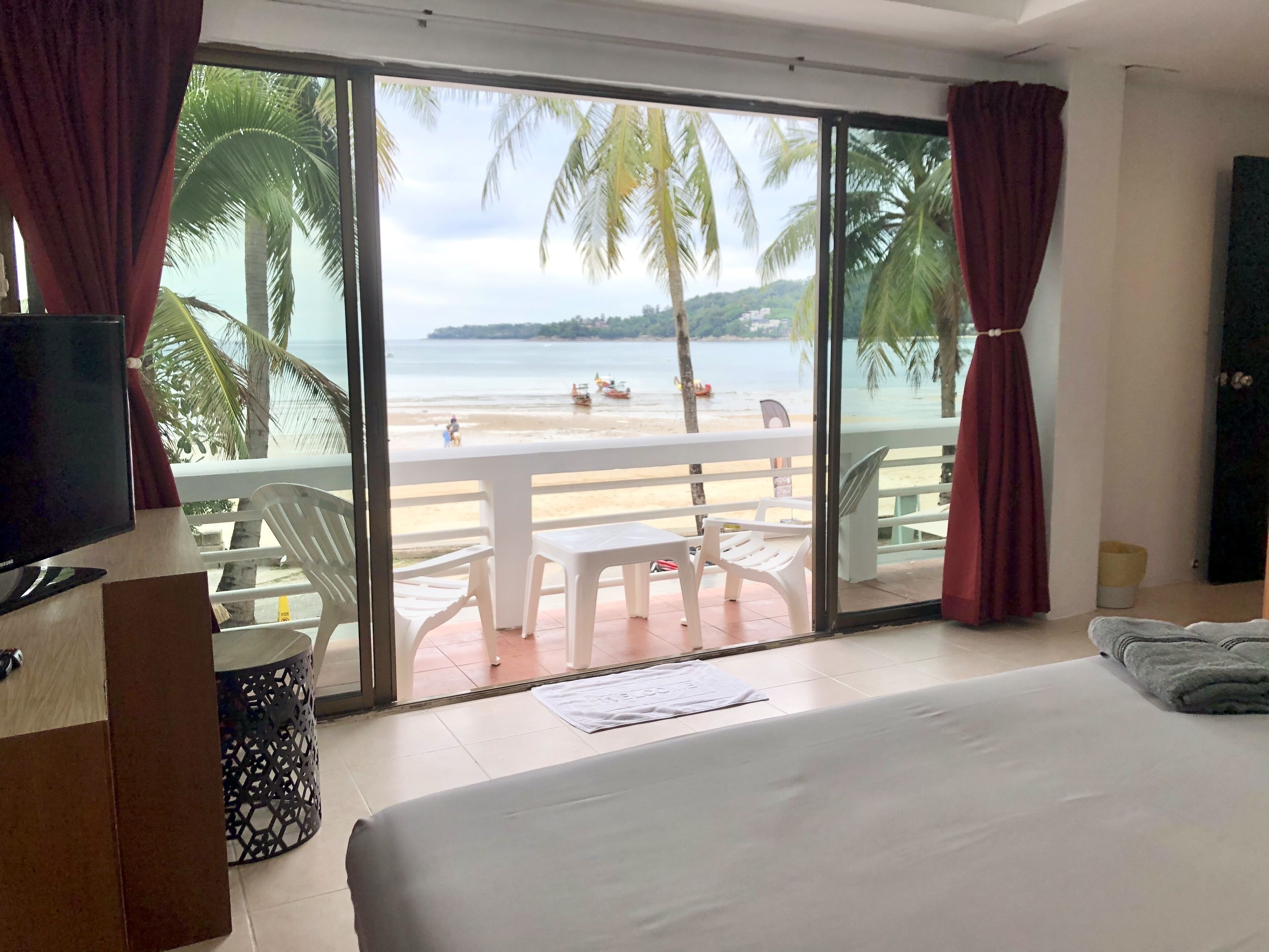 A room with a bed, a TV, and a view of a beach with palm trees.