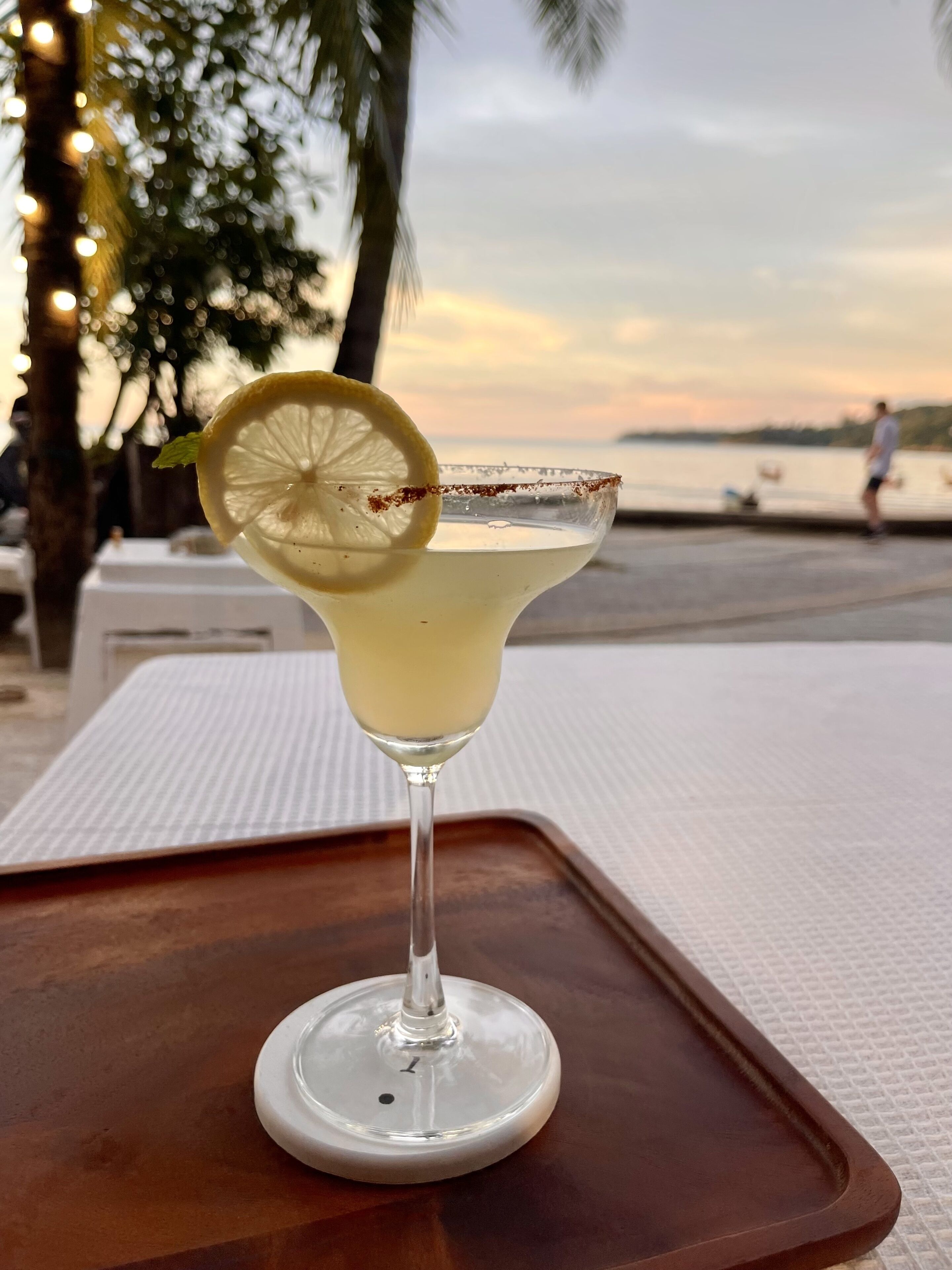 A cocktail with a lemon slice garnish on a white tray, set against a sunset backdrop.
