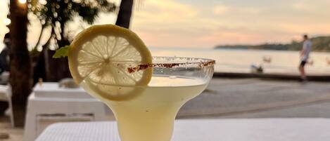 A cocktail with a lemon slice garnish on a white tray, set against a sunset backdrop.