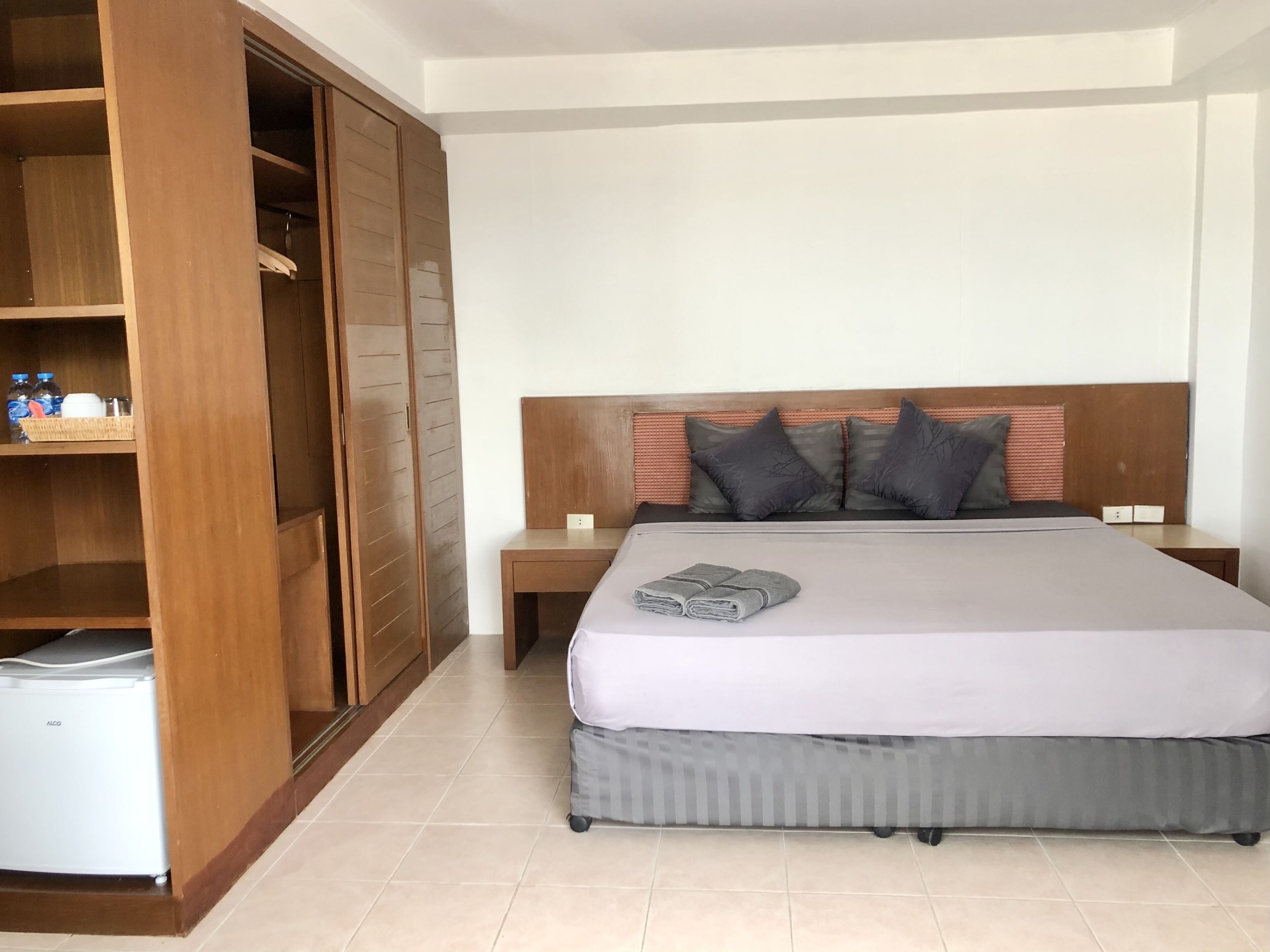 A hotel room with a large bed, a wooden wardrobe, a small bedside table, and a minibar.
