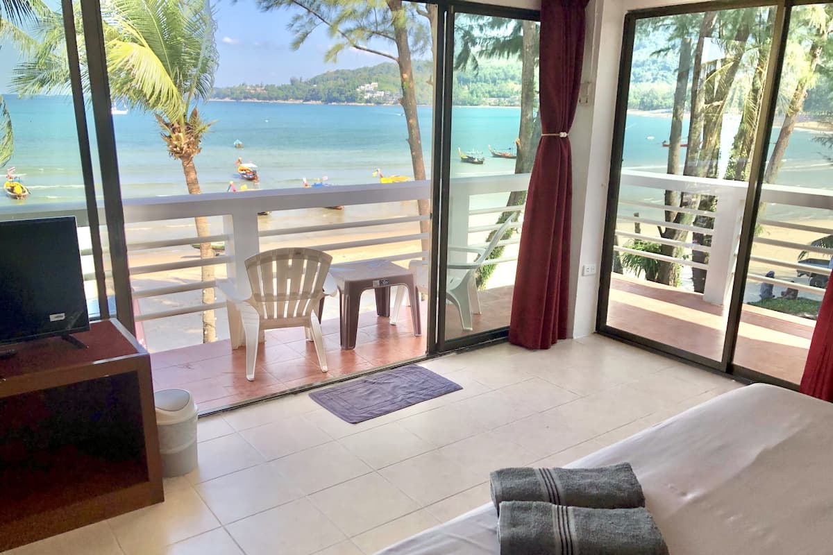 A hotel room with a bed, a TV, a balcony with a view of the sea and palm trees, and a chair.