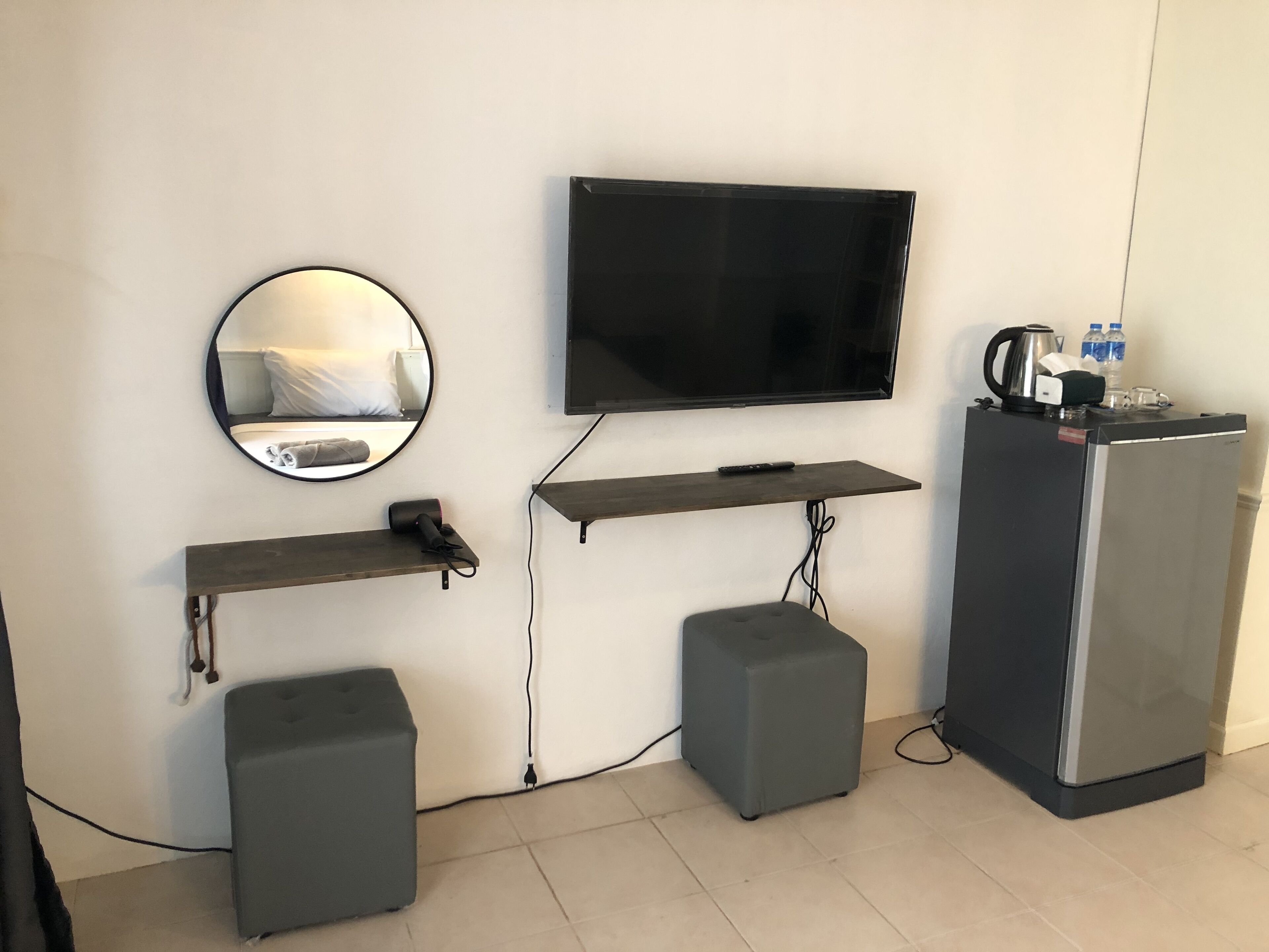 A hotel room with a flat-screen TV mounted on the wall, a round mirror, two grey ottomans, a small wooden shelf, and a refrigerator.