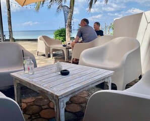 Un área de estar al aire libre con sillas de plástico blanco y una mesa de madera rústica, con vista a una playa con palmeras y el océano.