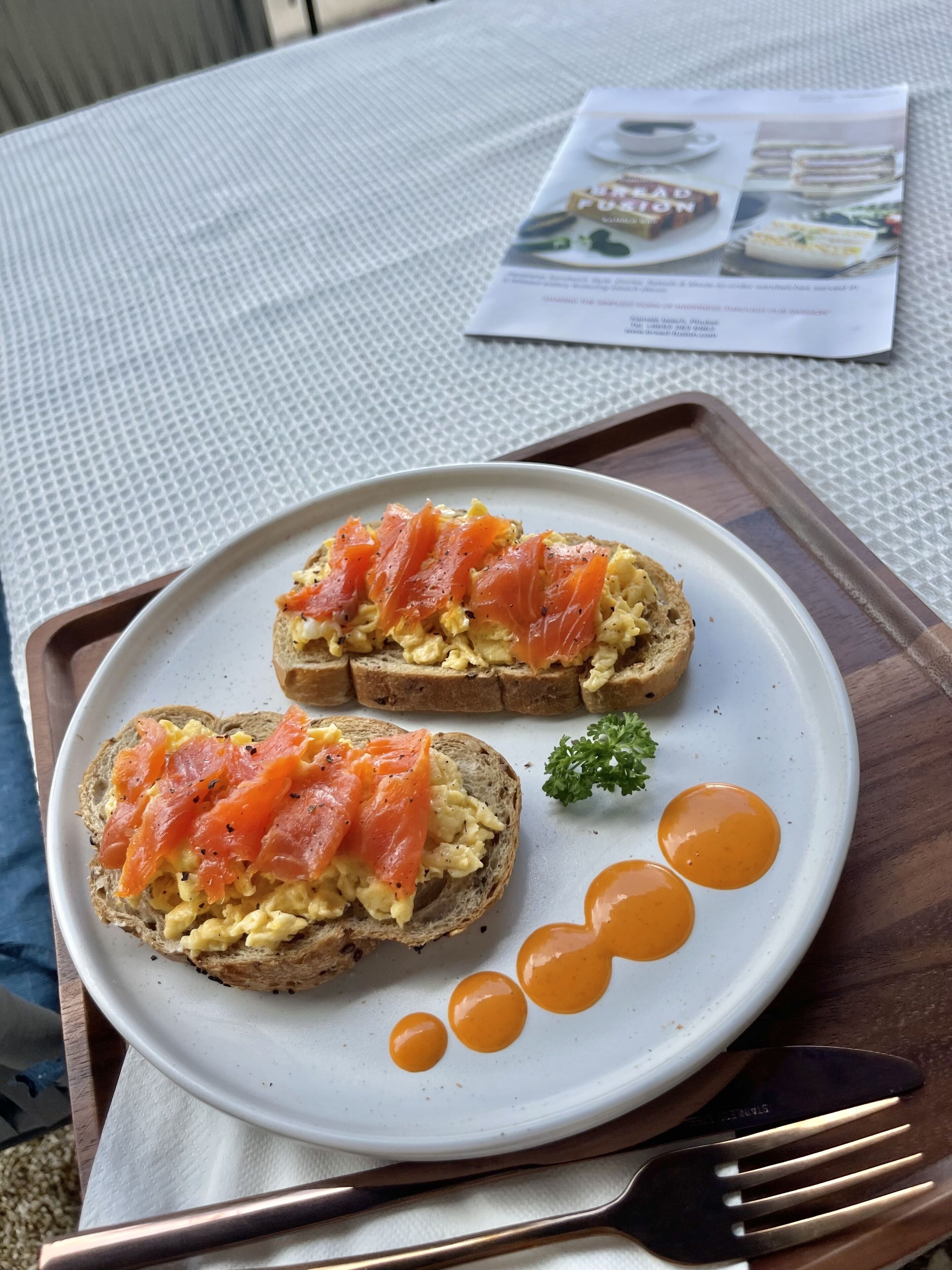 A plate with two open-faced sandwiches topped with smoked salmon and scrambled eggs, accompanied by a small sprig of parsley and three dollops of sauce.