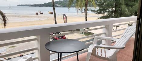 Varanda com cadeiras de plástico branco e uma mesa pequena, com vista para uma praia com palmeiras e uma linha de costa ao longe.