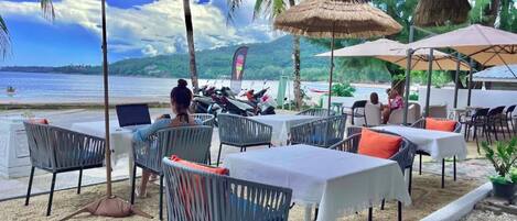 An outdoor seating area with tables and chairs, umbrellas, and a view of the beach and ocean.