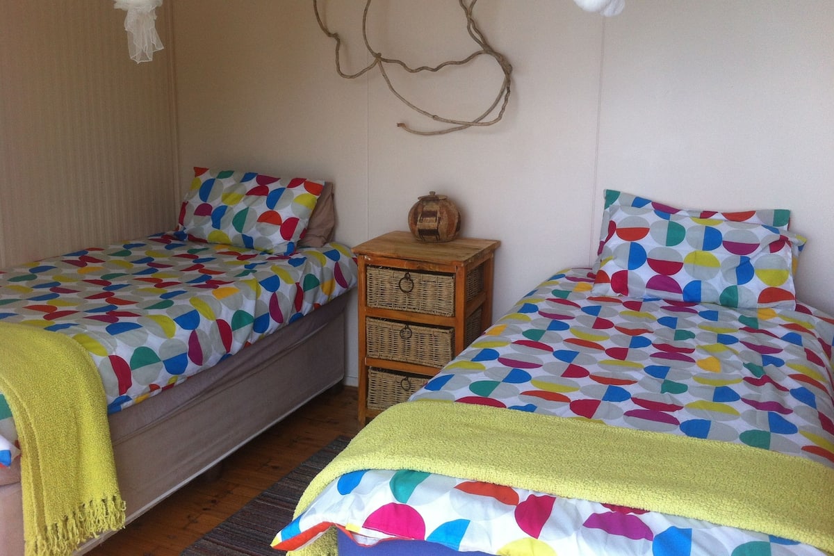 A bedroom with two single beds, each with colorful patterned bedding and a yellow throw blanket. There is a small wooden nightstand with a woven basket between the beds.