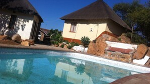 Una piscina amb vora enrajolada, envoltada de roques i plantes. Al fons s'hi veuen dos edificis amb teulades de palla.