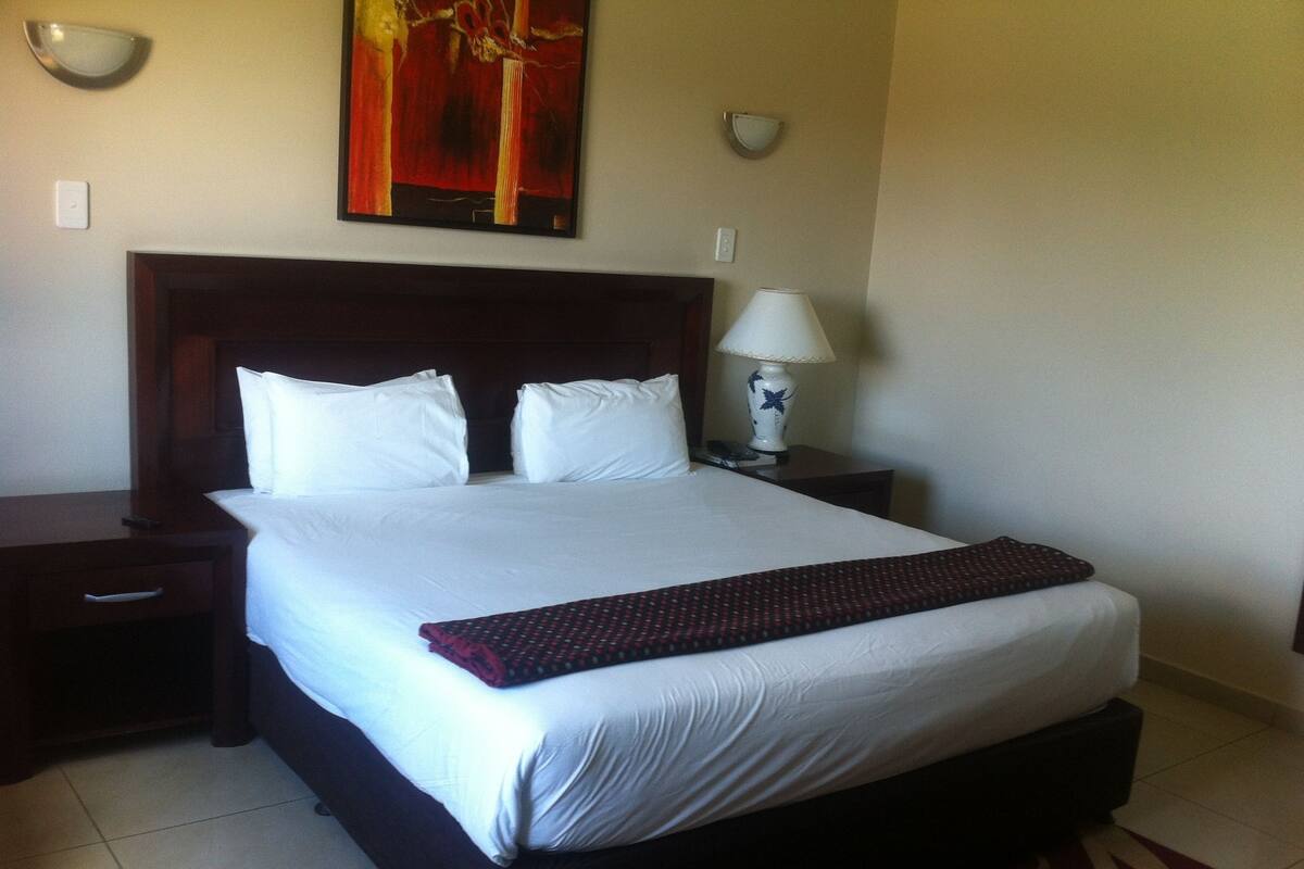 A neatly made bed with white linens, a dark headboard, and a red throw blanket. There is a white lamp on a bedside table and a painting above the bed.
