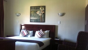 A hotel room with a large bed, a wooden headboard, two bedside tables, a chair, and a painting on the wall. - Desert Sands Motel (Palapye)