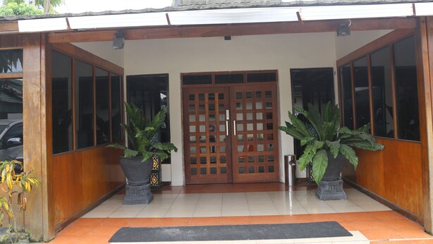 A building entrance with a tiled floor, a wooden door with glass panels, and two large potted plants. - Serayu Hotel Timika (Timika)