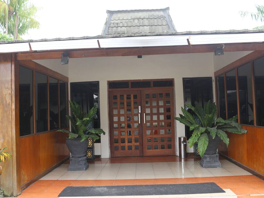 A building entrance with a tiled floor, a wooden door with glass panels, and two large potted plants.