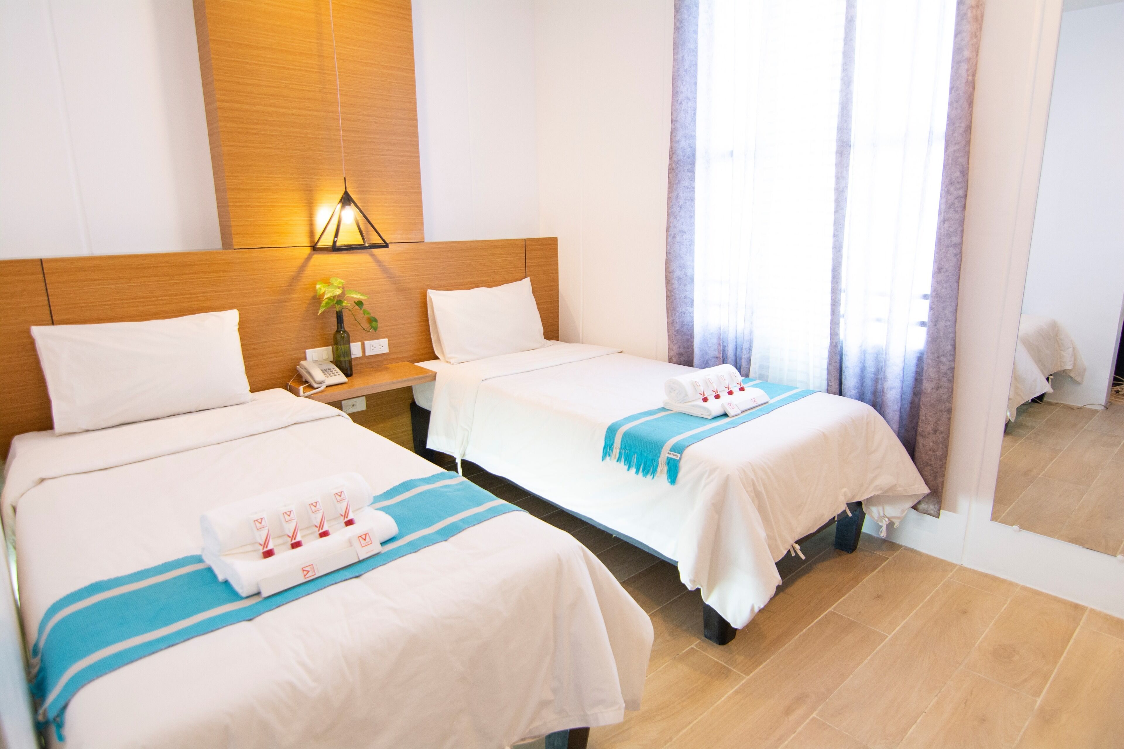 Habitació d'hotel amb dos llits, un panell de fusta al capçal, una làmpada penjada a la paret, un telèfon i una finestra amb cortines.