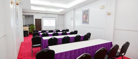 Un salón de conferencias con alfombra roja, paredes blancas y manteles morados. Hay sillas negras alrededor de las mesas, y se ve una pantalla de proyector al fondo.