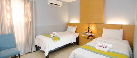 A hotel room with two beds, a chair, an air conditioning unit, and a bedside table with a lamp.