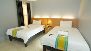 A hotel room with two single beds, each with a white bedspread and a colorful throw blanket. There is a nightstand with a lamp between the beds, and a window with curtains on the left side. - Viven Hotel (San Nicolas)