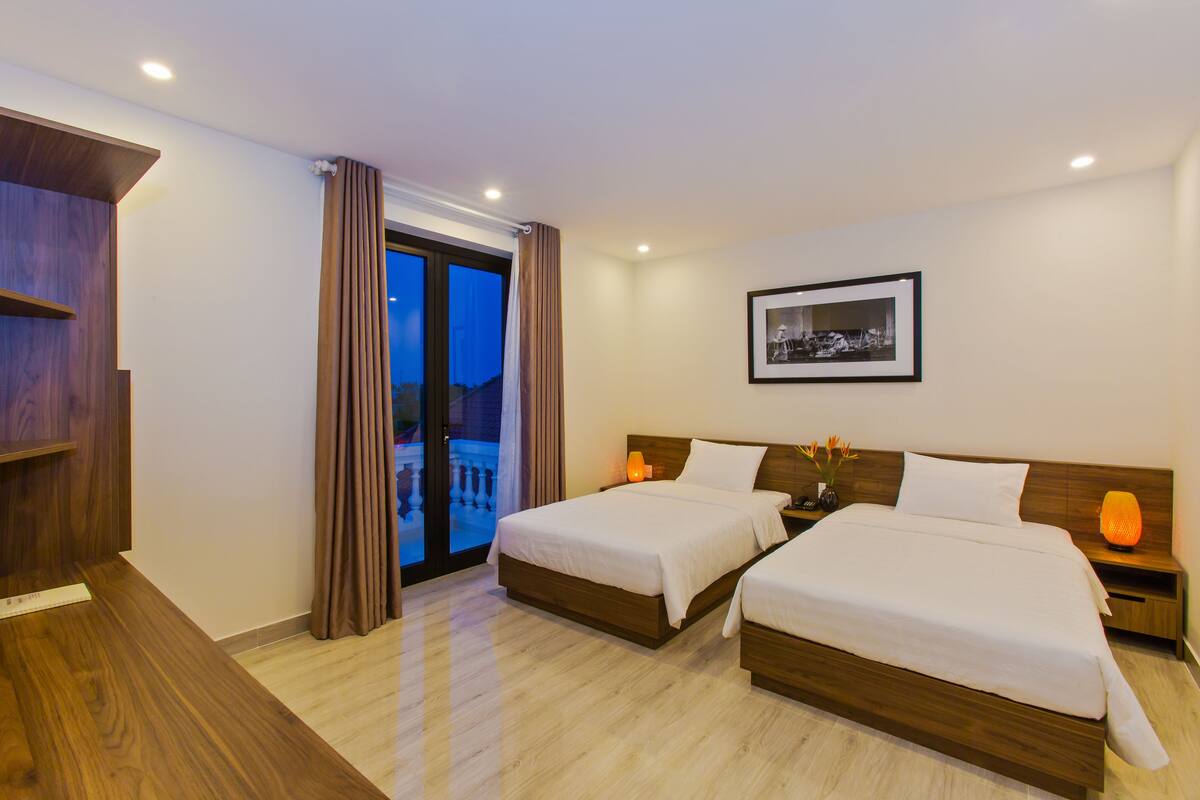 A hotel room with two beds, a wooden desk, a nightstand with a lamp, and a framed picture on the wall.