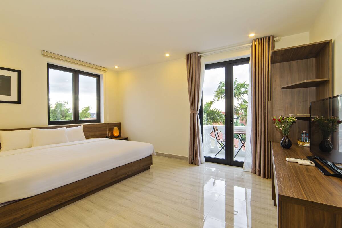 A hotel room with a large bed, a TV, a desk with a vase of flowers, and a balcony with a view of palm trees.