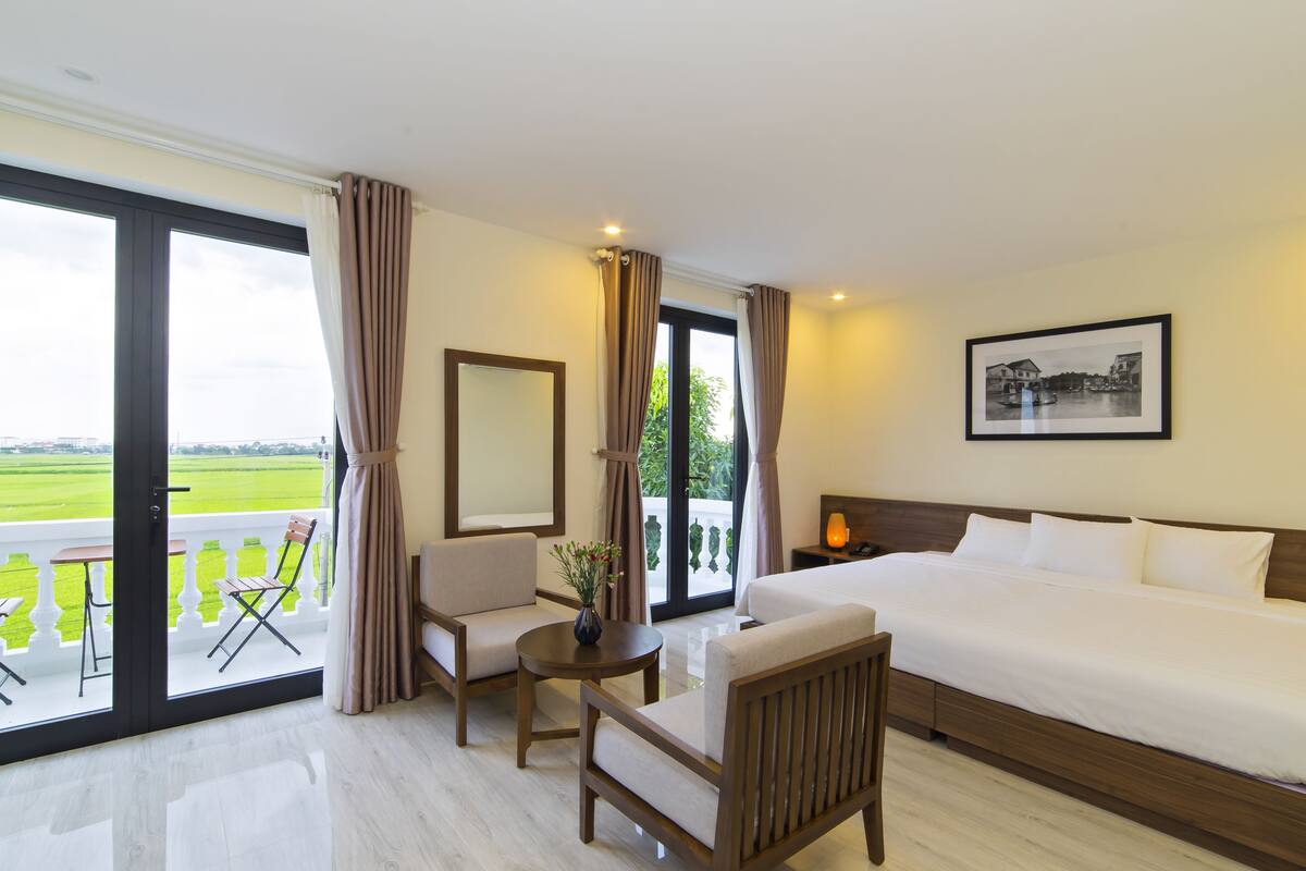 A hotel room with a large bed, a chair, a small table, a mirror, and a view of the outdoors.