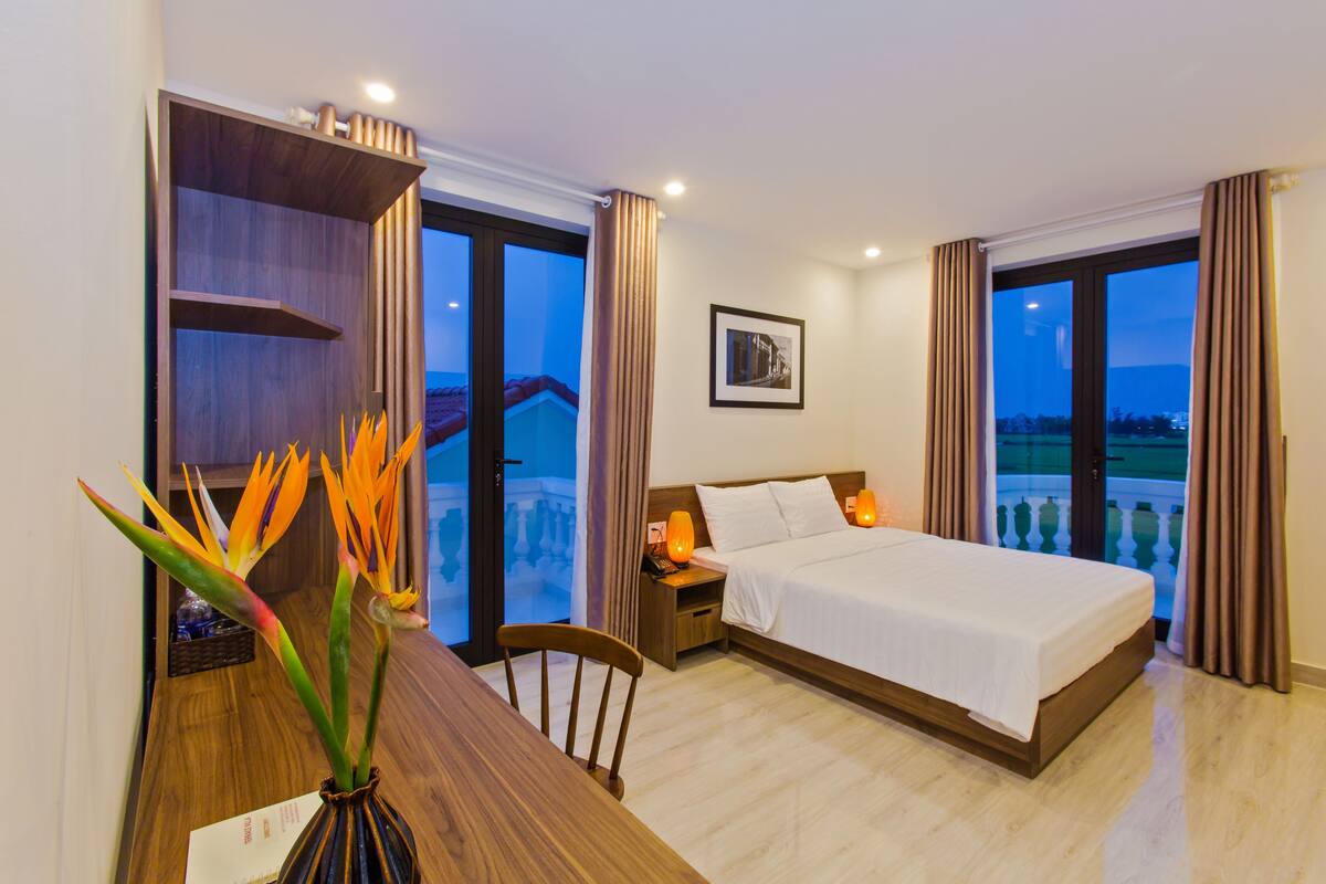 A hotel room with a bed, a desk, a chair, a nightstand, a lamp, a picture on the wall, and a view of the outside.