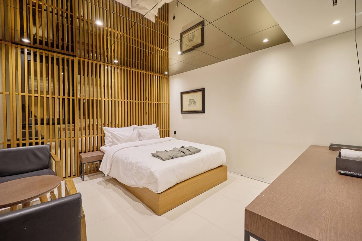 A modern hotel room with a wooden bed, a small bedside table, a sofa, and a wooden desk.