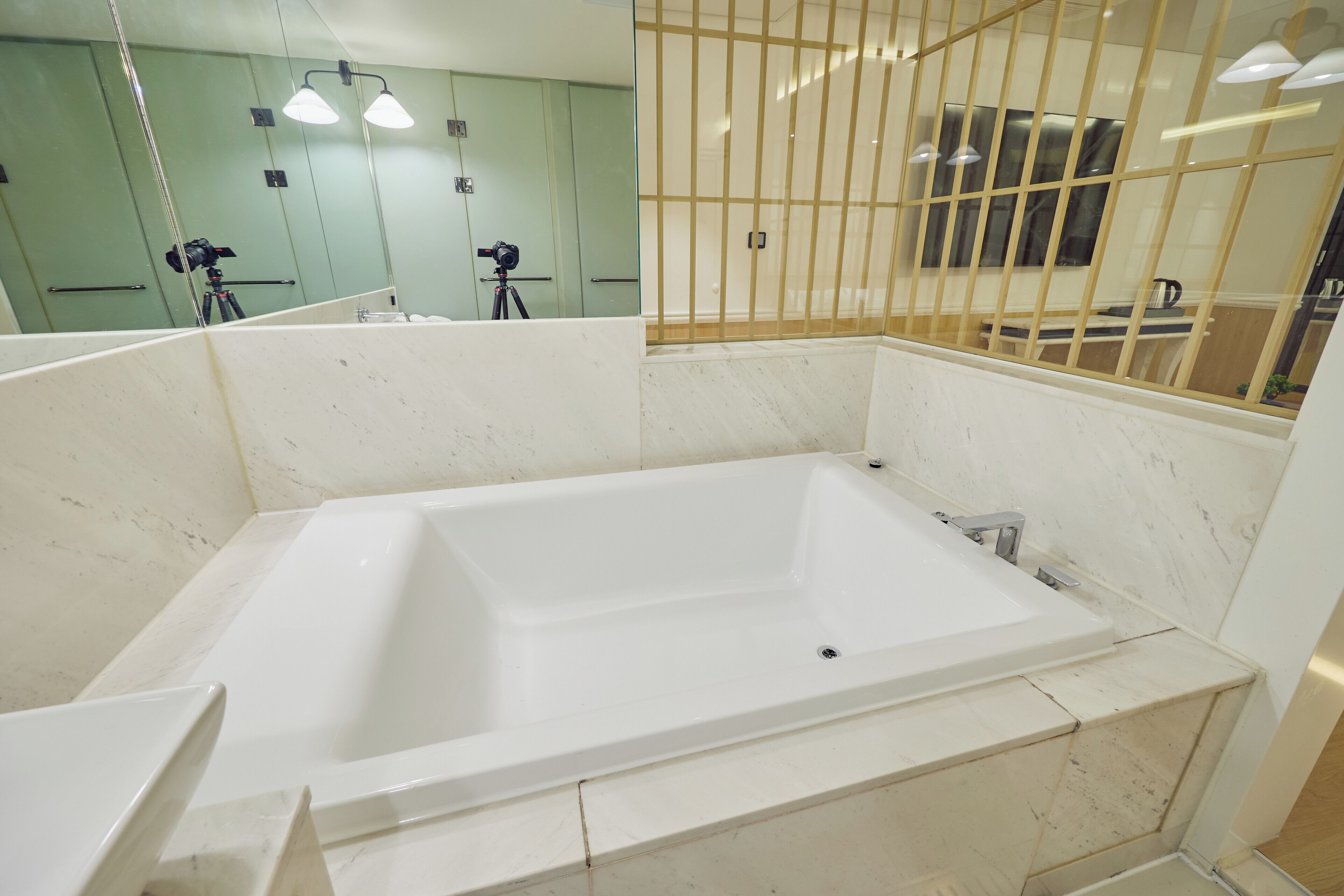 A modern bathroom with a large rectangular bathtub, a mirror, and a camera on a tripod in the background.