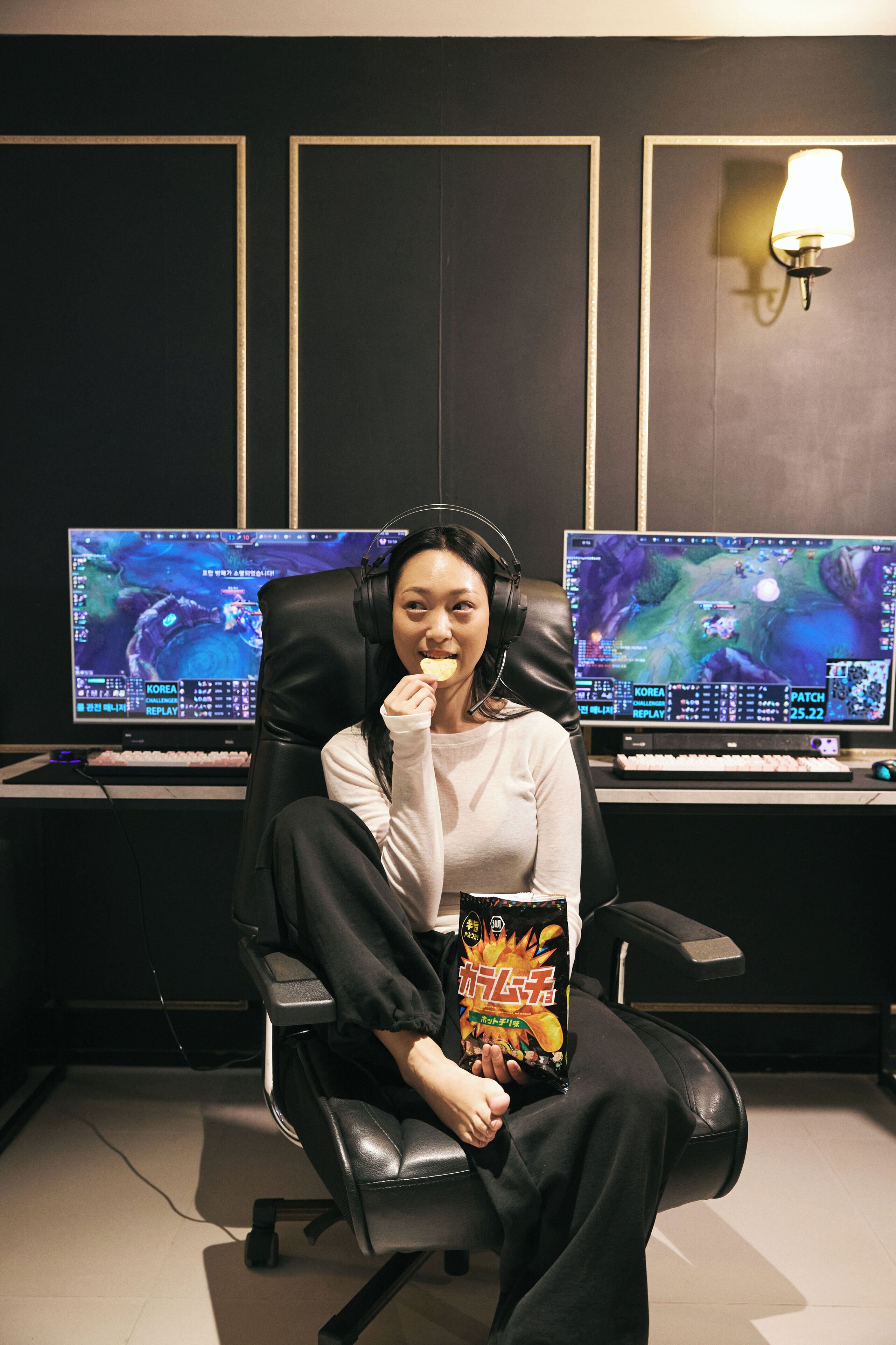Eine Person sitzt in einem Stuhl, im Hintergrund ist eine Gaming-Station mit zwei Monitoren zu sehen, die ein Videospiel zeigen.