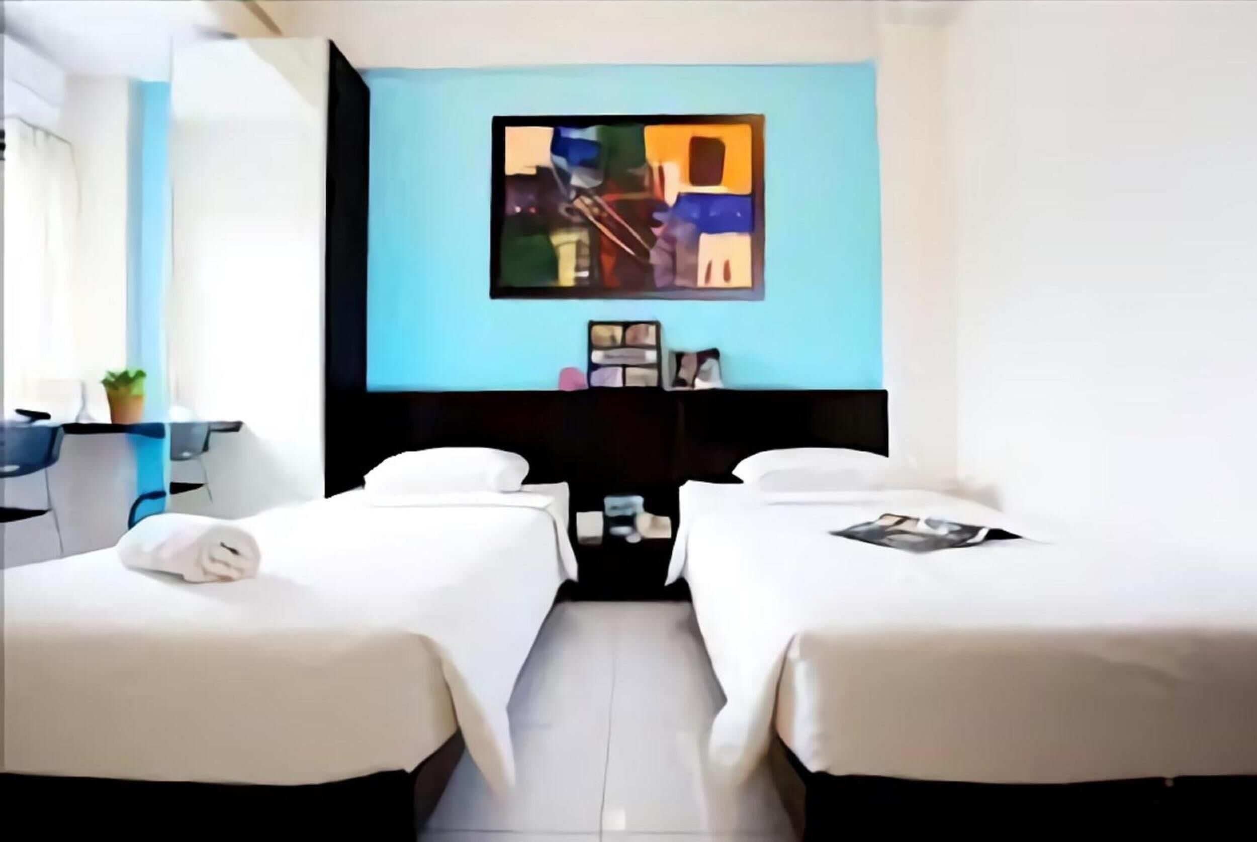 Habitación de hotel con dos camas, una pared de acento azul, un cuadro y un pequeño escritorio con una planta.