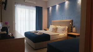 A hotel room with a bed, a desk, and a chair. There is a window with curtains, an air conditioning unit, and a nightstand with a lamp.