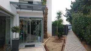 A modern building with a glass entrance, a paved pathway, and a small garden area with plants and a palm tree.