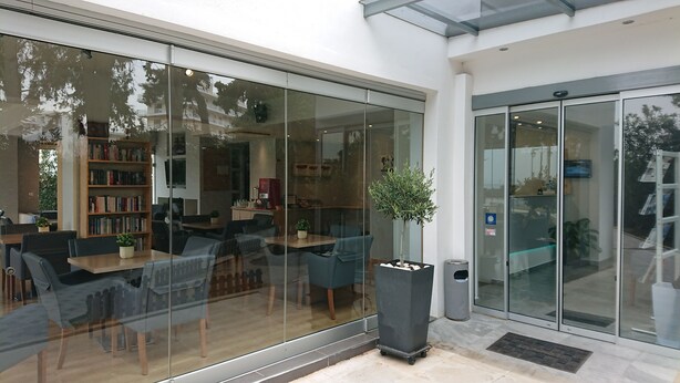 A modern indoor space with glass walls, a seating area with tables and chairs, a bookshelf, and a trash bin.