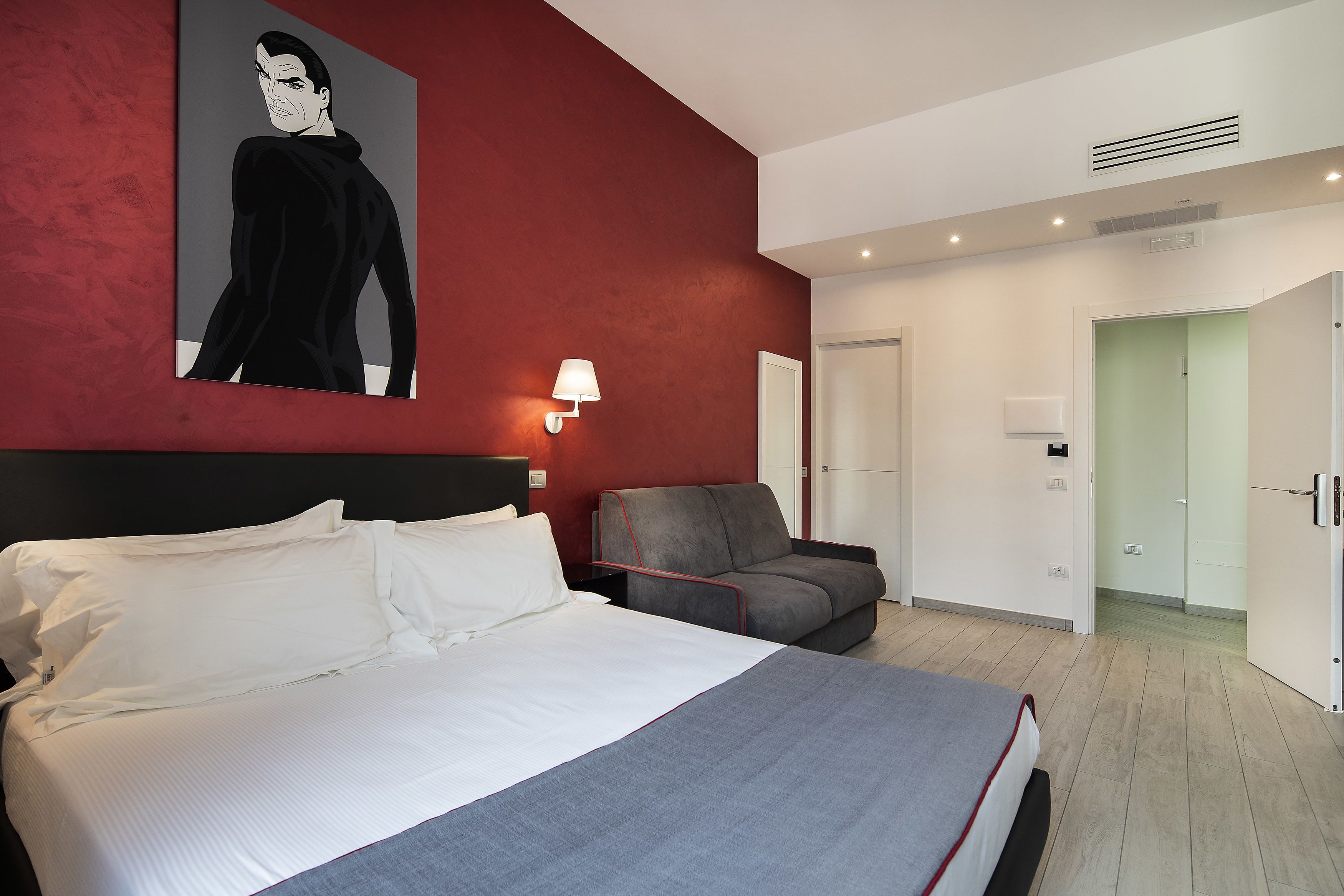 Une chambre d’hôtel avec un grand lit, un canapé gris, un mur d’accent rouge et un tableau représentant un homme.