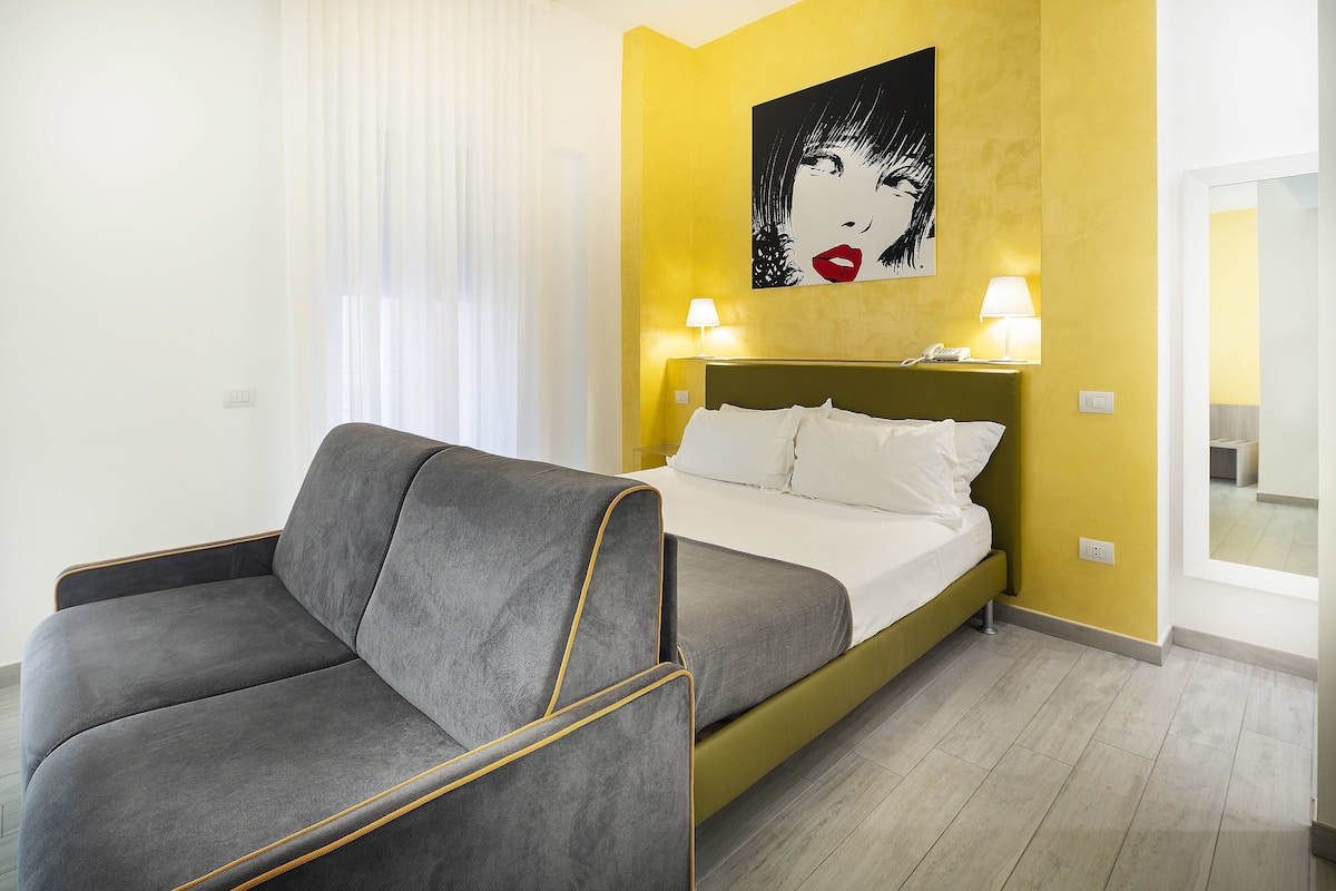 Een hotelkamer met een bed, een grijze bank, twee bedlampen en een groot schilderij aan de muur.