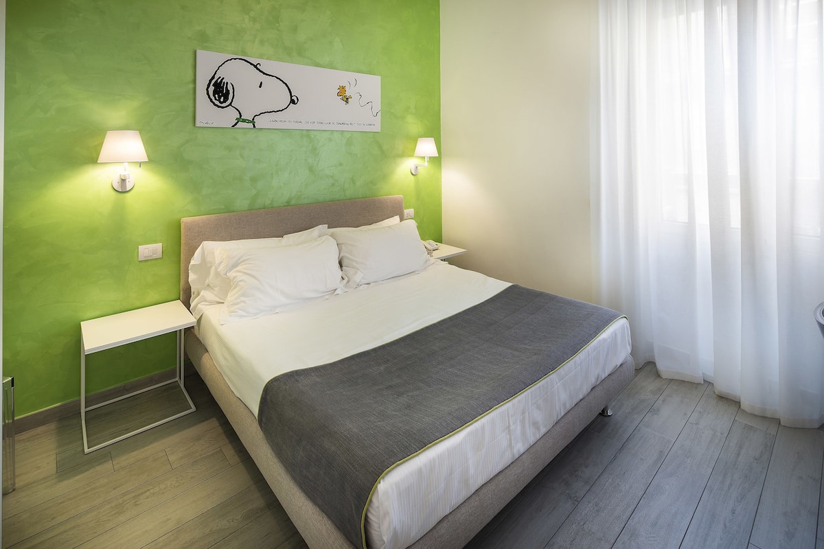 Een netjes opgemaakt bed met een grijze deken, twee nachtkastjes met lampen, een groene wand en een raam met dunne gordijnen.
