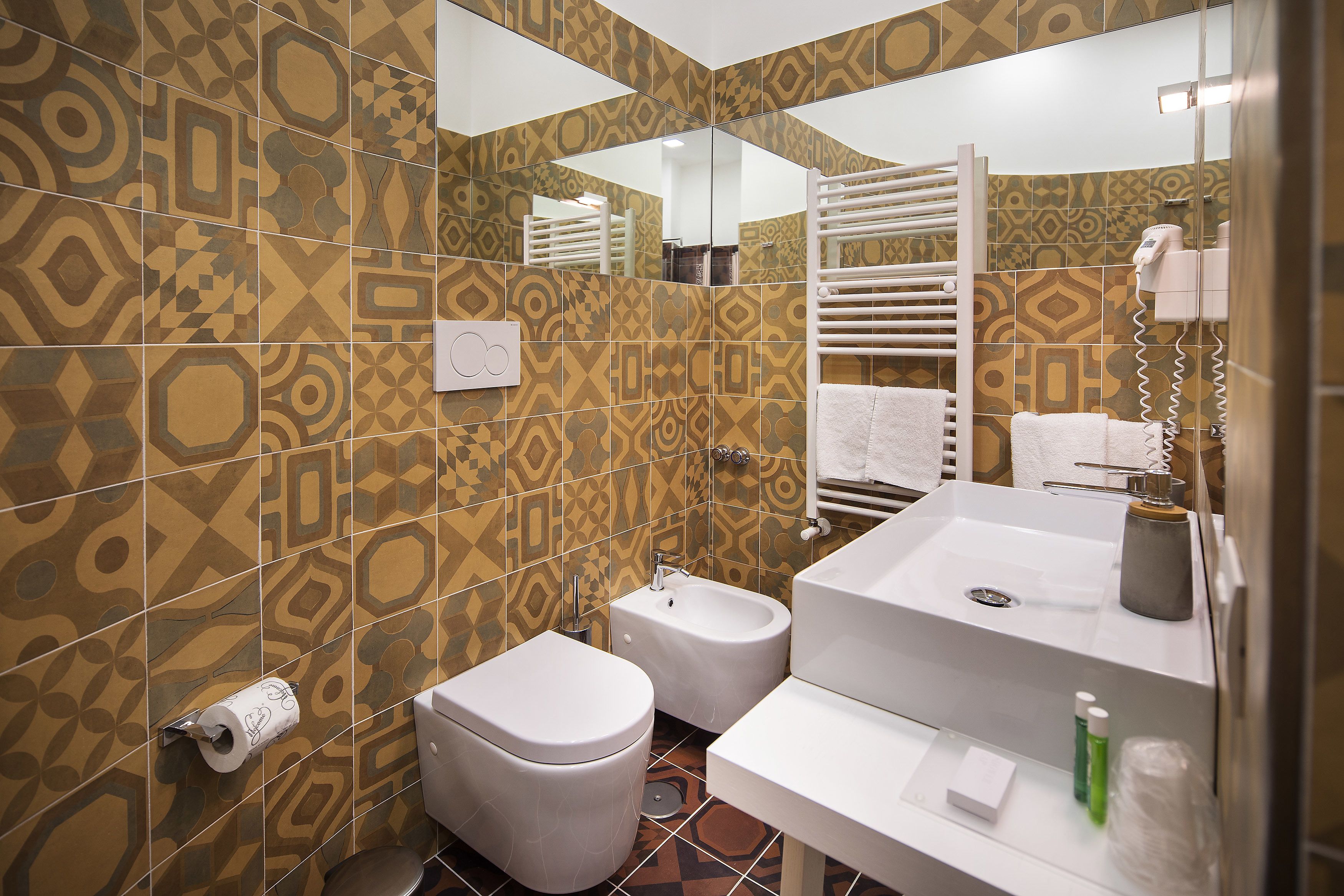 A bathroom with patterned tiles, a toilet, bidet, sink, towel rack, and a mirror.