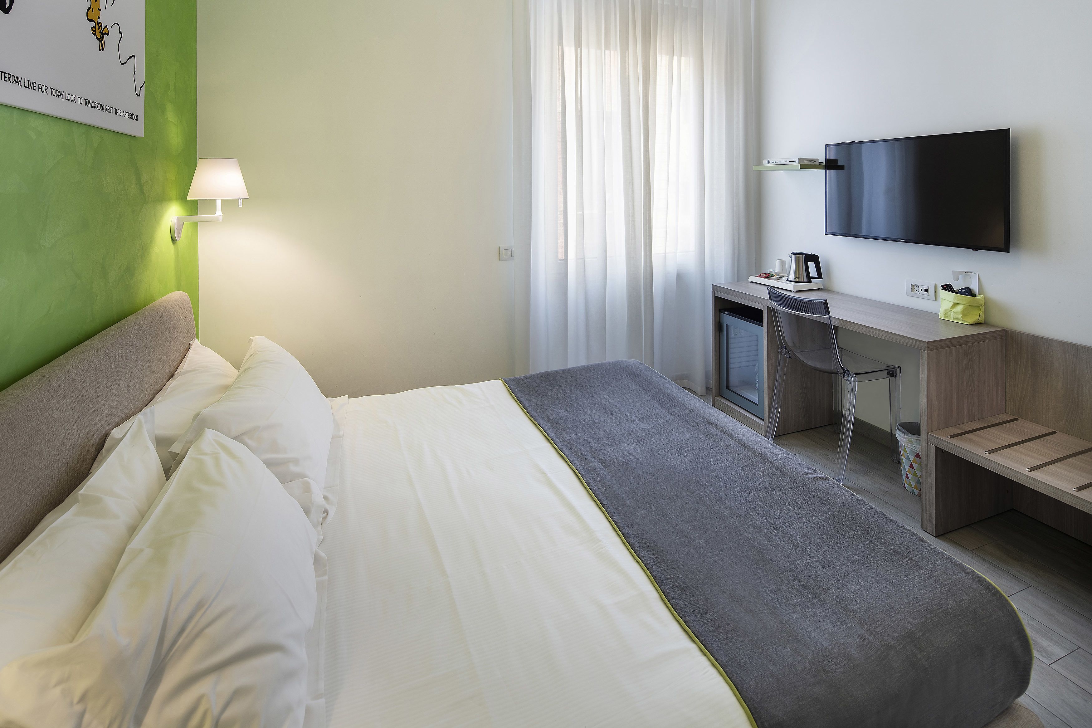 A hotel room with a bed, a desk with a chair, a flat-screen TV, a lamp, and a window with sheer curtains.
