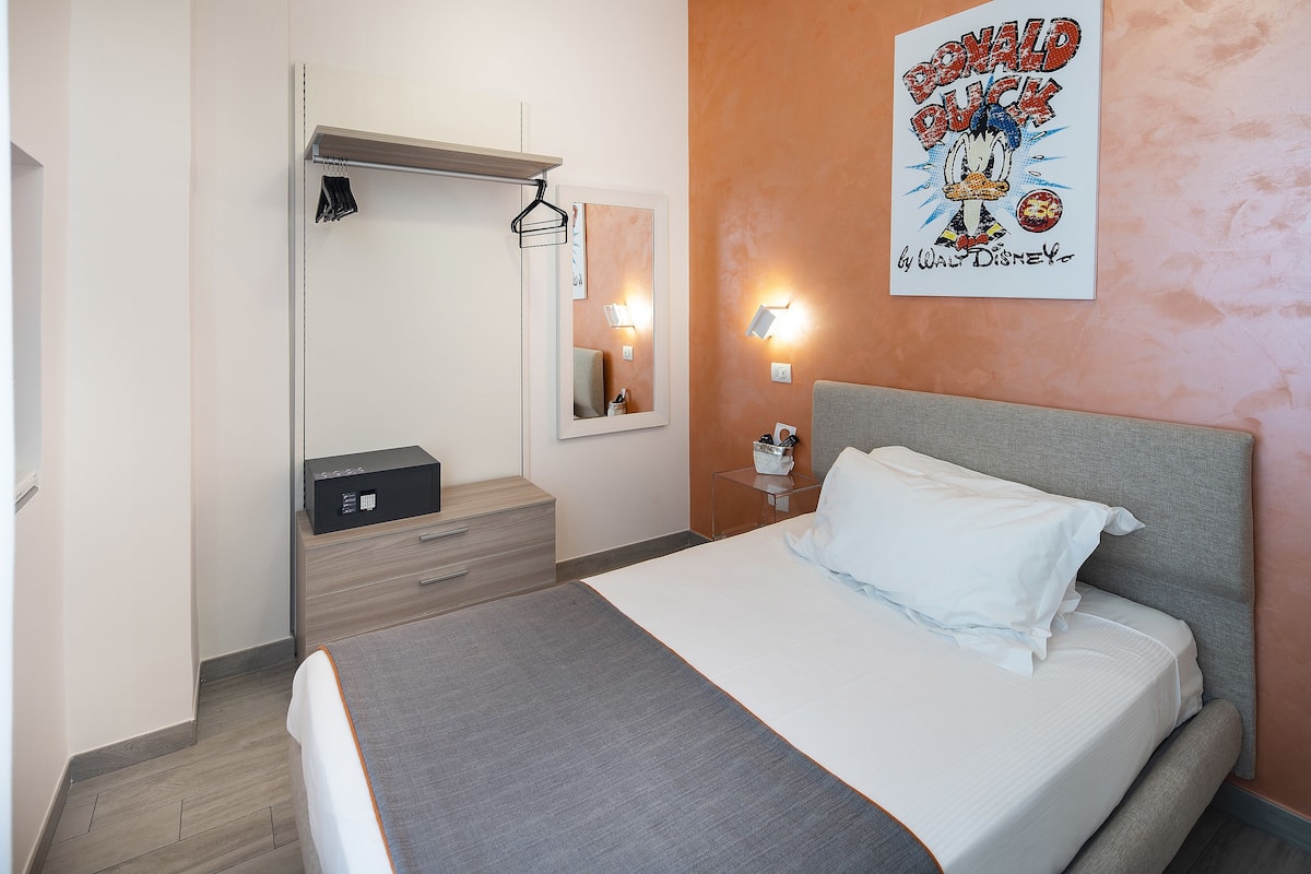 Een netjes opgemaakte hotelkamer met een bed, nachtkastje, lamp en een poster van Donald Duck.
