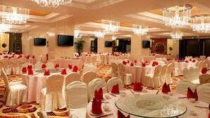 Un salón de banquetes con mesas redondas, sillas blancas, servilletas rojas y varios candelabros.