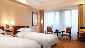 Habitación de hotel con dos camas, una zona de estar con una silla y una mesita, y un ventanal grande con cortinas.