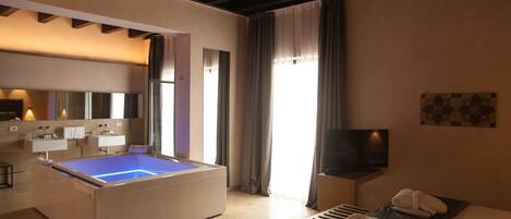 Habitación de hotel moderna con una bañera grande, televisor de pantalla plana, cama con toallas y baño con espejo y lavamanos.