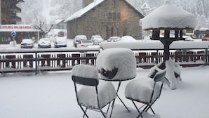 A snowy outdoor seating area with a thatched roof structure, a stone building with a clock tower, and parked cars.