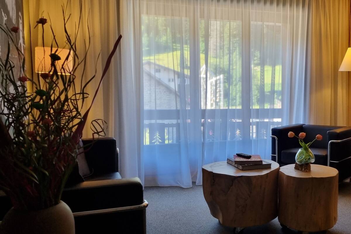 A cozy living room with a sofa, two round wooden side tables, a vase with flowers, a fireplace, and a view of a wooden deck and greenery outside.