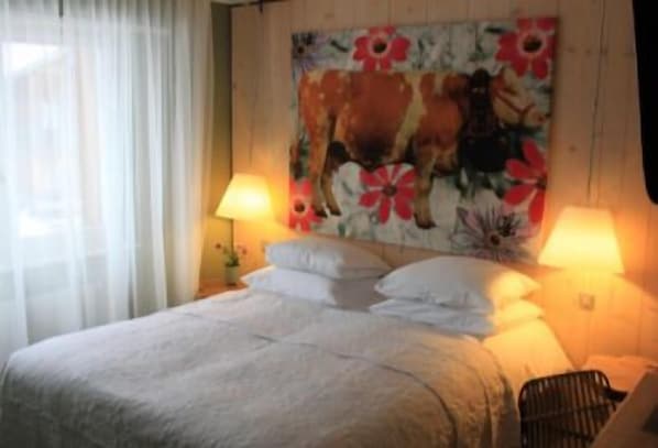 Un dormitorio con cama, dos lámparas de noche, un cuadro floral en la pared y una ventana con cortinas.